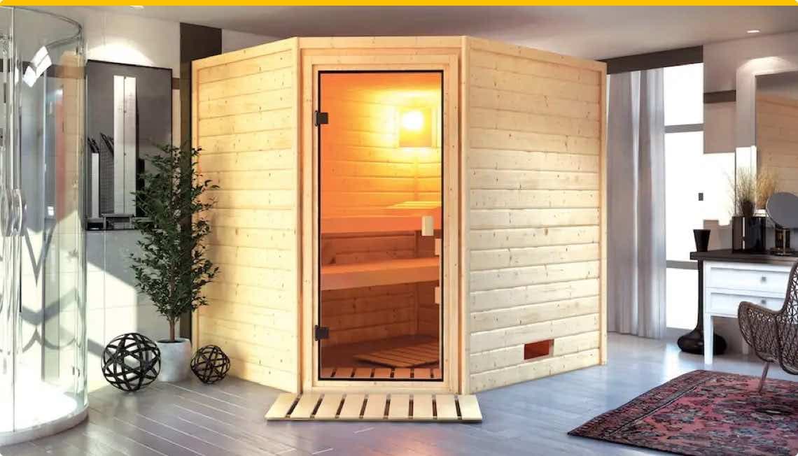 Holzsauna mit geöffneter Glastür in modernem Zimmer mit Teppich und Pflanzen, neben Dusche und Schreibtisch mit Stuhl
