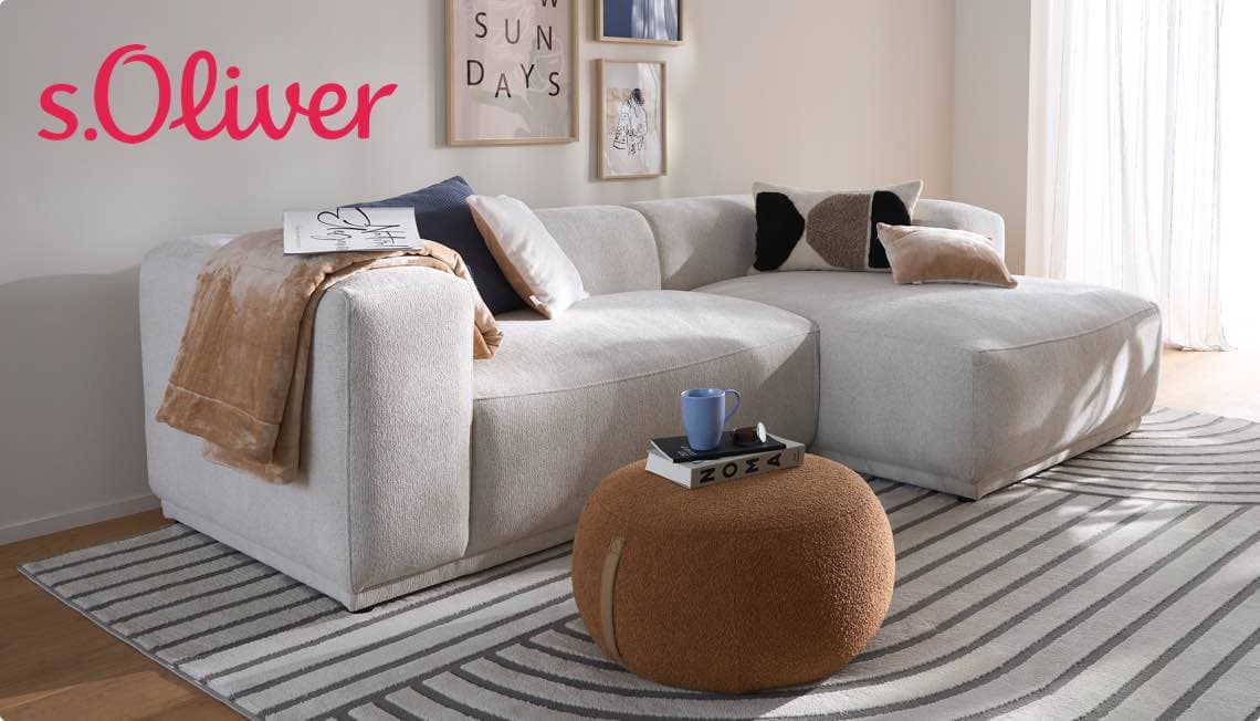 s.Oliver – gemütliches Wohnzimmer mit hellgrauem Ecksofa, brauner Decke, bunten Kissen und rundem Hocker mit Buch und Tasse