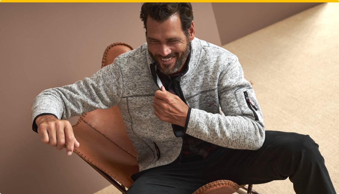 Mann sitzt auf einem Lederstuhl und trägt eine graue Jacke mit schwarzen Details.