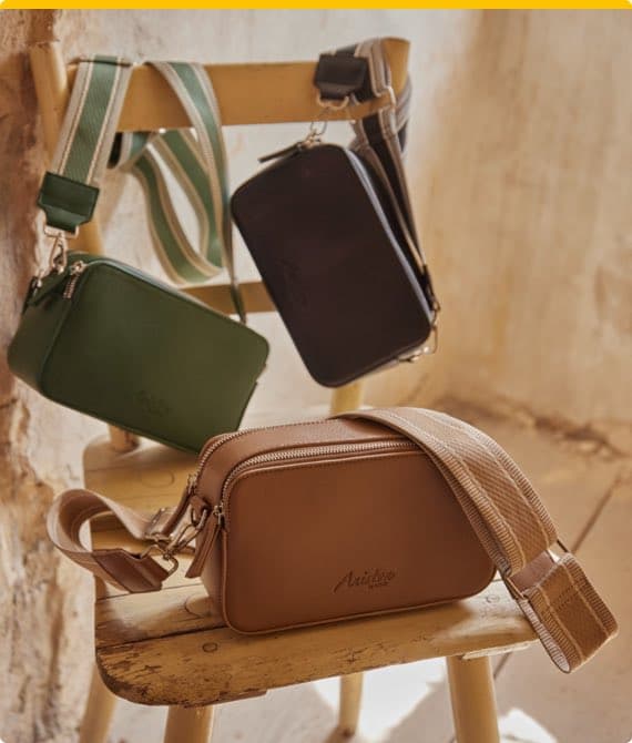 Drei Umhängetaschen in Grün, Schwarz und Beige hängen und liegen auf einem Stuhl vor einer Wand.