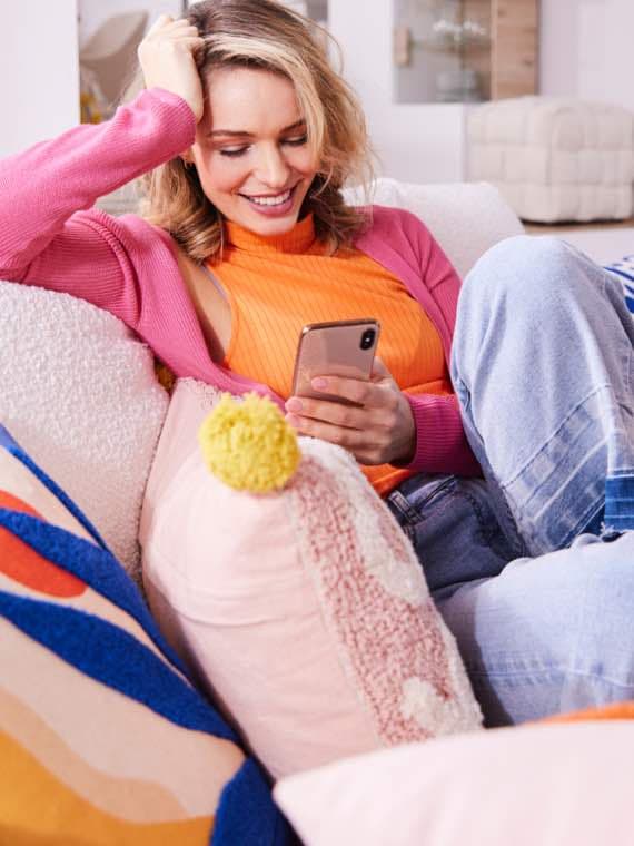 Person in pink Pullover und orangefarbenem Top sitzt entspannt auf dem Sofa und schaut auf ihr Handy.