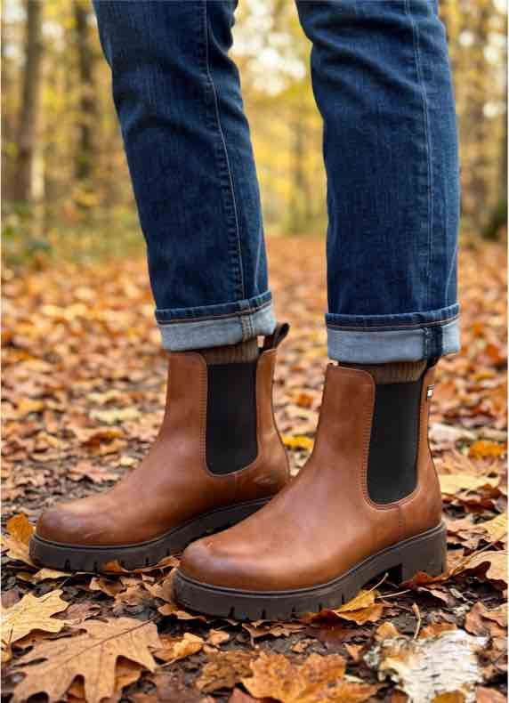 Person in hochgekrempelter Jeans trägt braune Stiefeletten und steht auf herbstlichem Laubweg im Wald